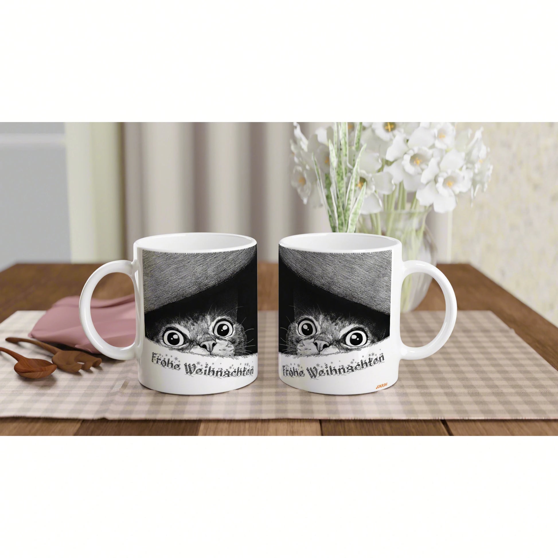 Two white mugs with a cat-face peeking from beneath a curved black stripe; 'Frohe Weihnachten' on each mug.