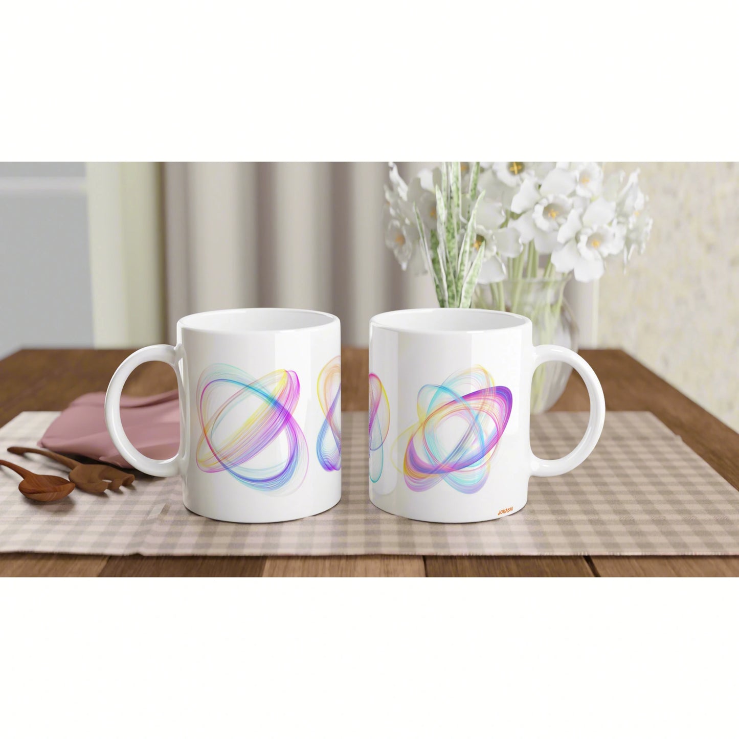 Two white mugs with colorful abstract swirl patterns sit on a wooden table with a checkered placemat and a vase of white flowers in the background.