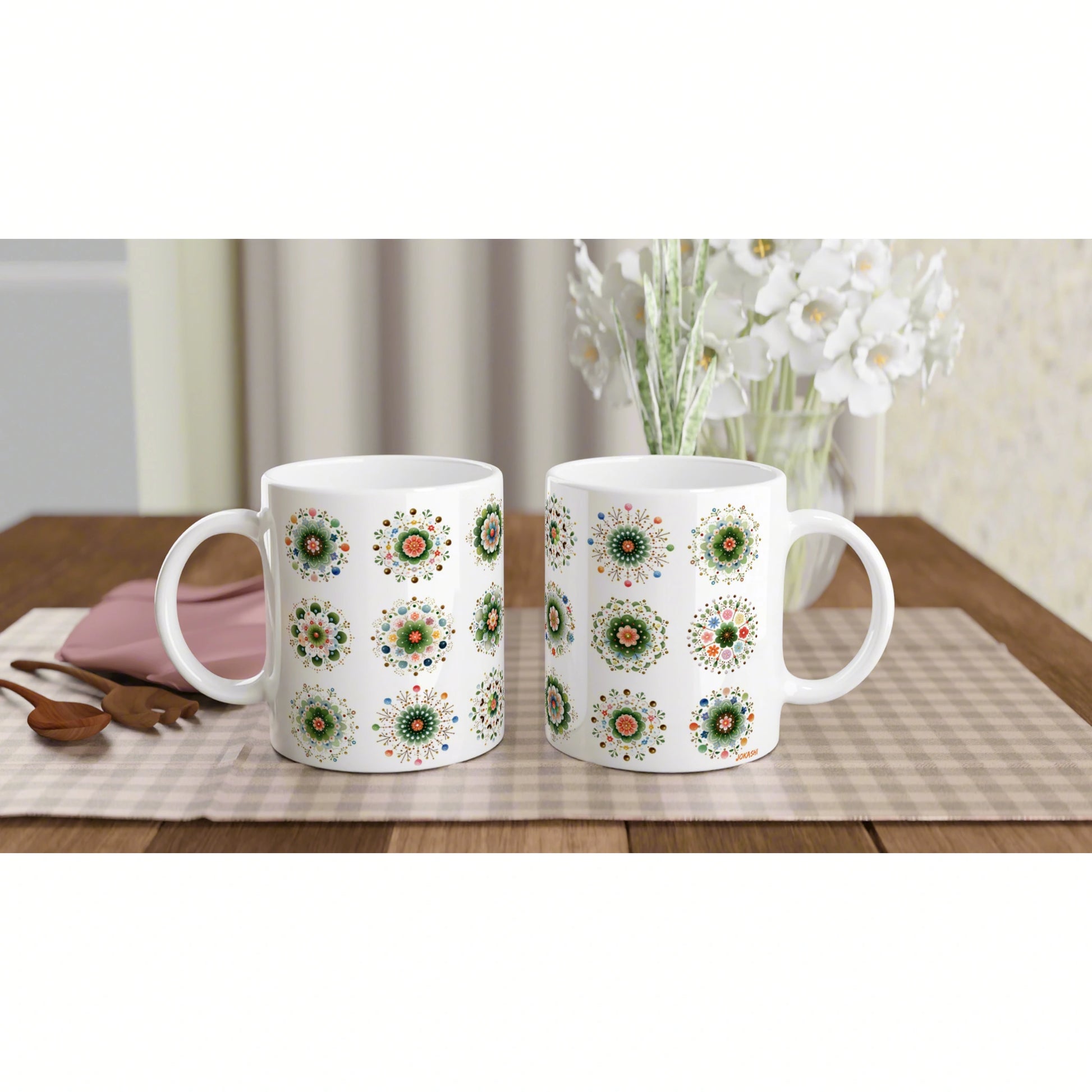 Two white ceramic mugs with colorful circular floral patterns sit on a wooden table with a beige placemat; white flowers in the background.