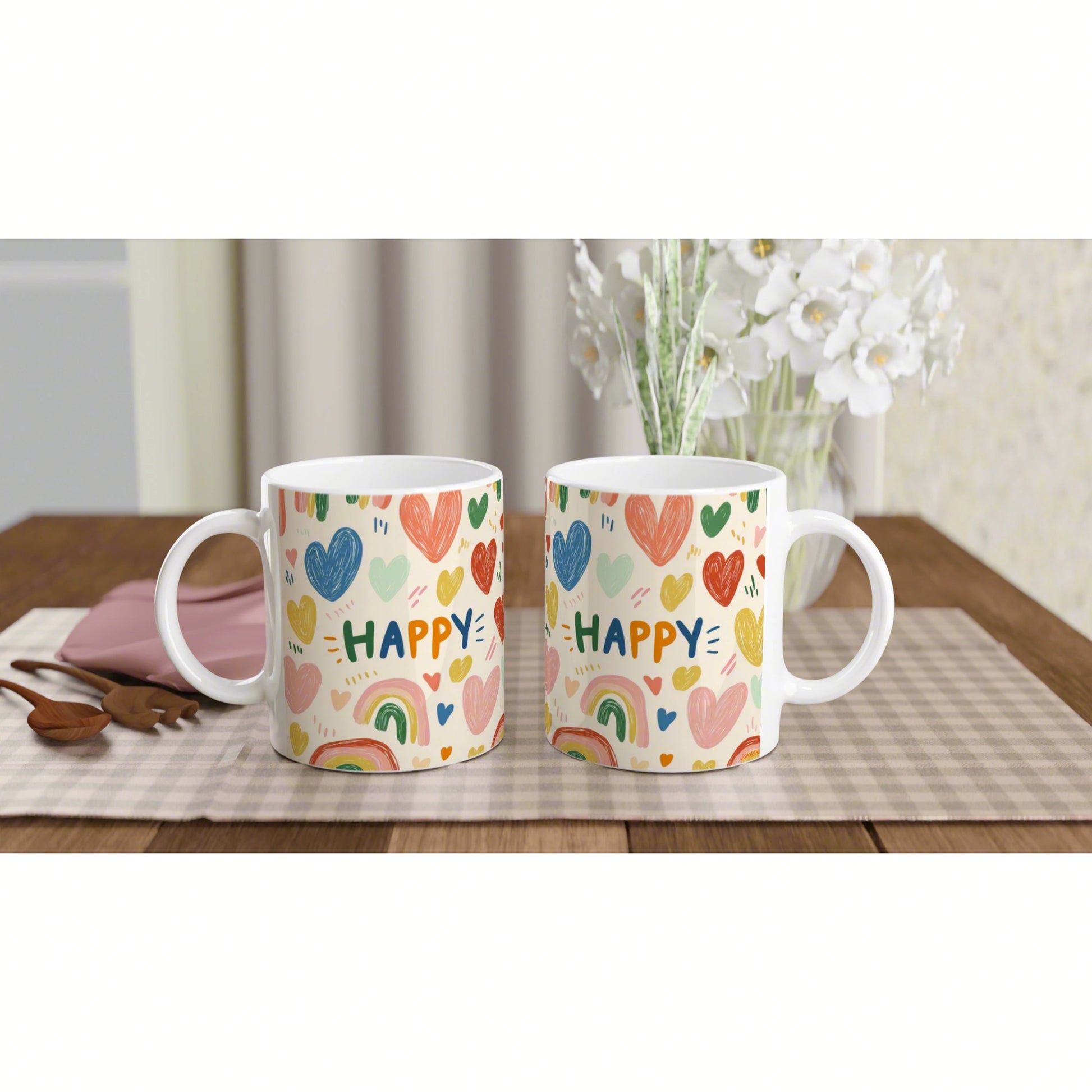 Two white mugs with colorful hearts and rainbows spelling HAPPY, on a checkered table runner with a vase of white flowers.
