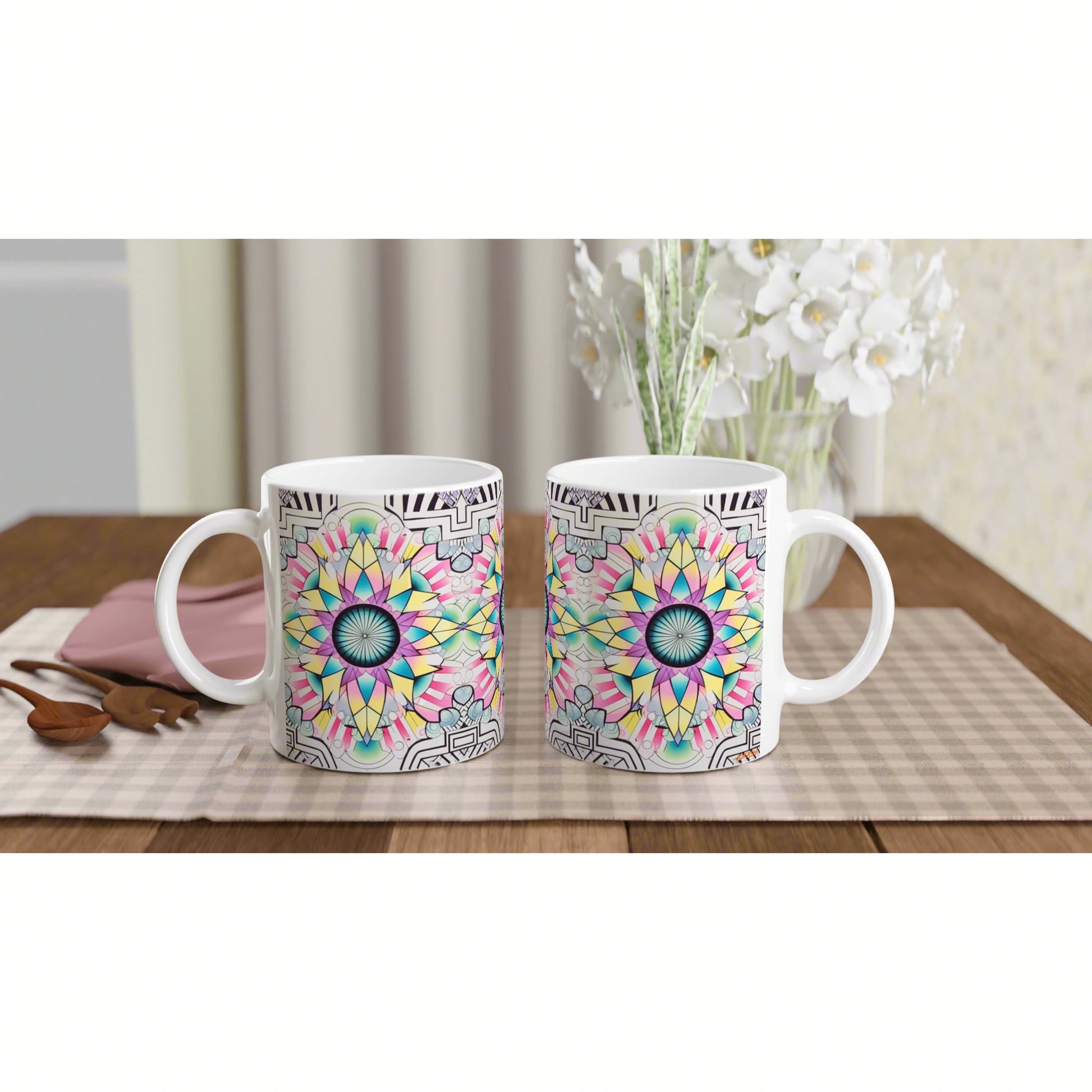 Two white mugs with colorful mandala patterns sit on a checkered placemat on a wooden table, with a vase of white flowers.