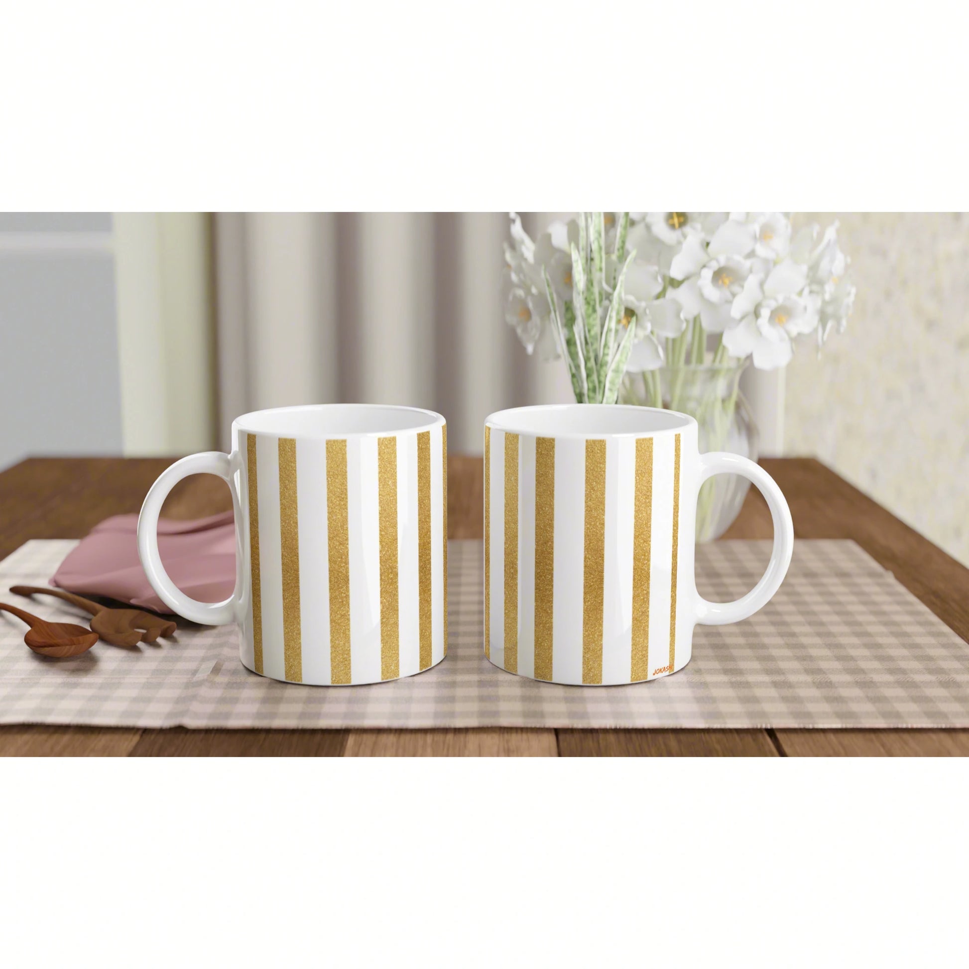 Two white mugs with gold vertical stripes sit on a plaid placemat on a wooden table; background shows a vase of white flowers.
