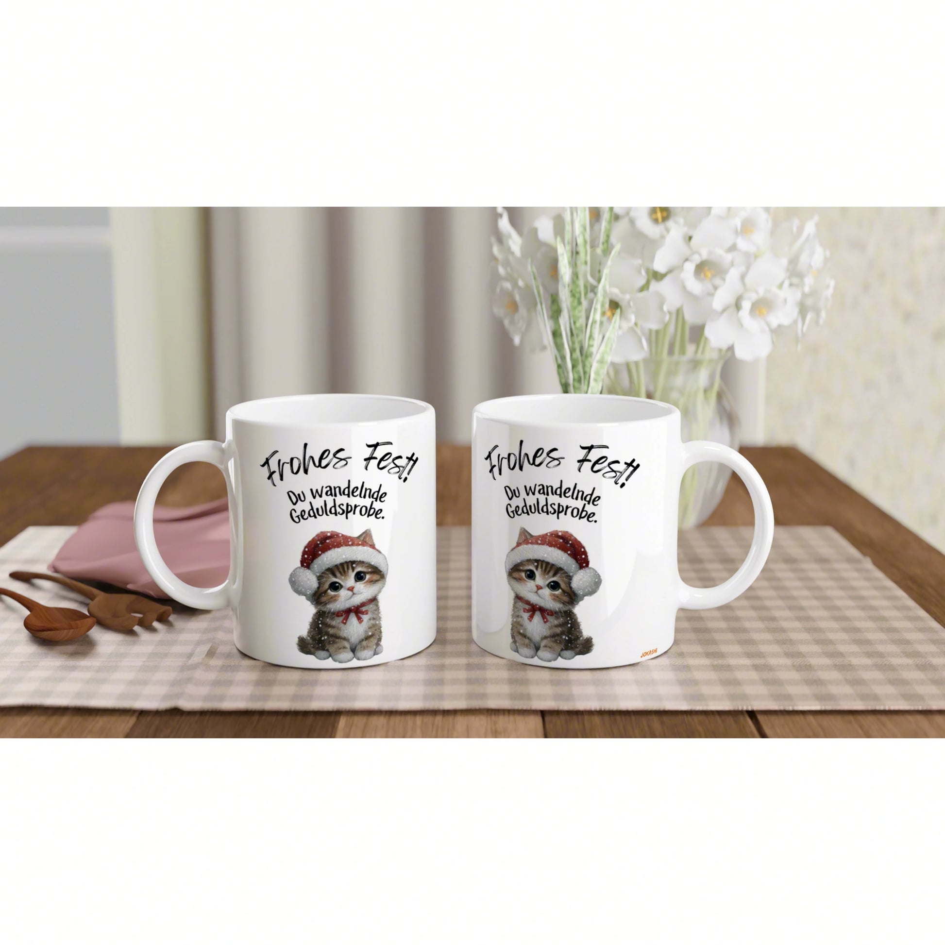 Product mockup: two white mugs with a kitten in a Santa hat and 'Frohes Fest', on a checkered table runner.