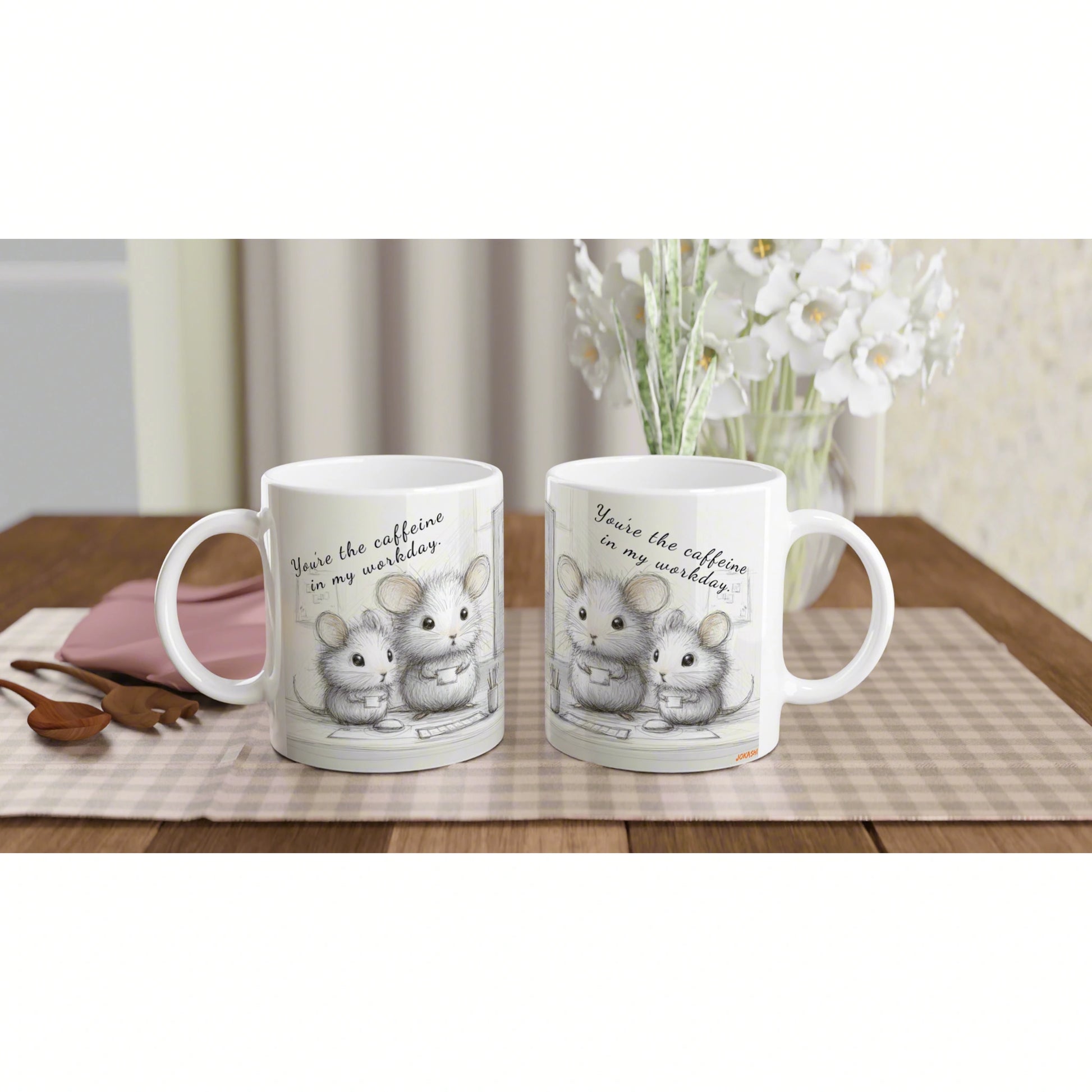 Two white mugs with a cute mouse illustration and the slogan 'You're the caffeine in my workday' on a checkered placemat.