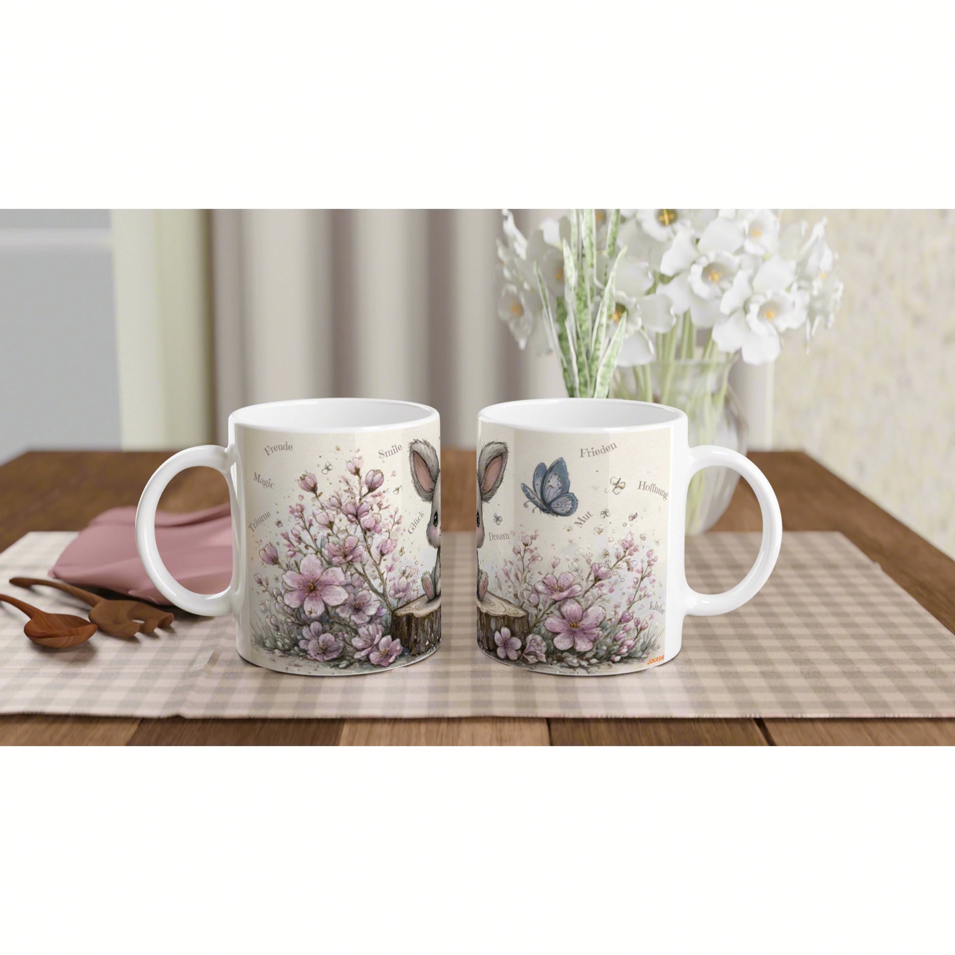Two white mugs with pastel floral and butterfly designs on a checkered placemat, on a wooden table with white flowers behind.