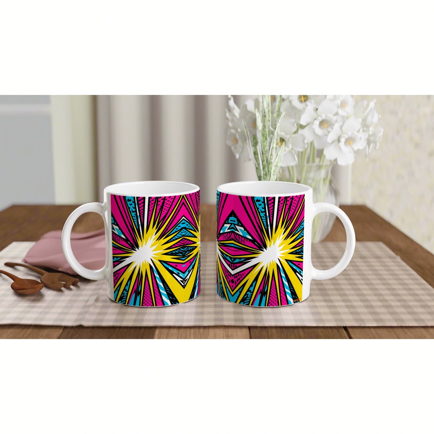 Two white mugs with pink, yellow and cyan starburst pattern on a checkered placemat, with white flowers in the background.