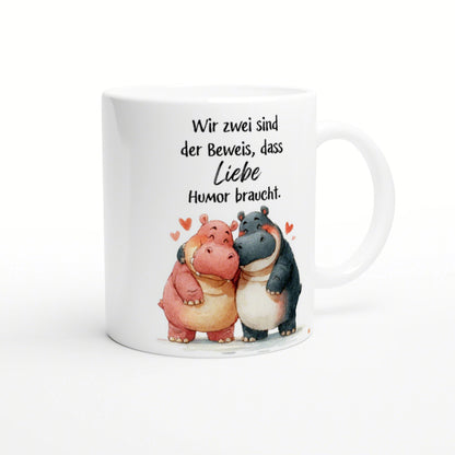 Weiße Kaffeetasse mit zwei cartoon-Nilpferden, die sich umarmen, und dem Spruch 'Liebe braucht Humor'.