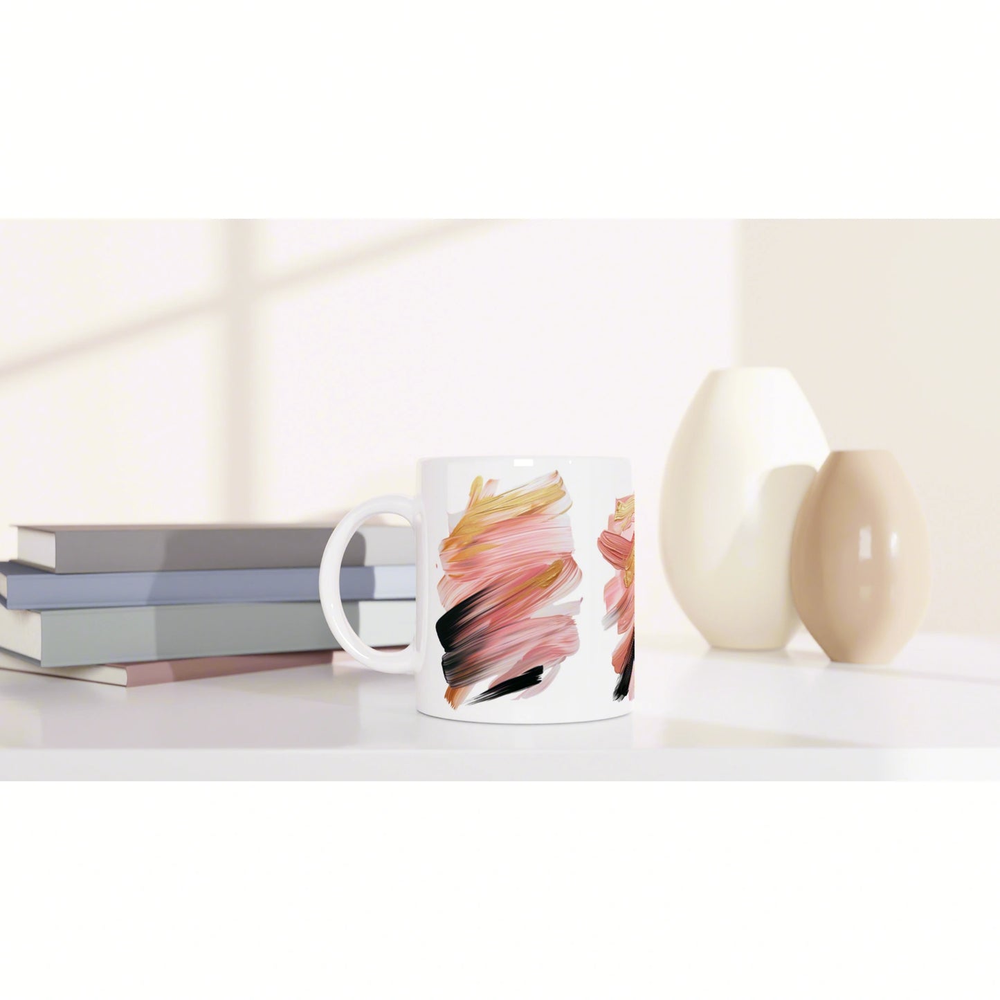 White ceramic mug with colorful abstract brushstrokes, beside stacked books and pale ceramic vases on a clean surface.
