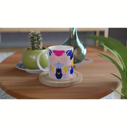 White ceramic mug with bright abstract multicolor design on a wooden coaster, on a table beside a green cactus.