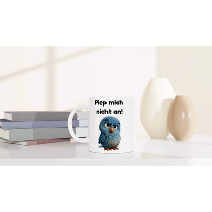 White ceramic mug featuring a blue cartoon bird and the text 'Piep mich nicht an!' on a clean desk with books and vases.