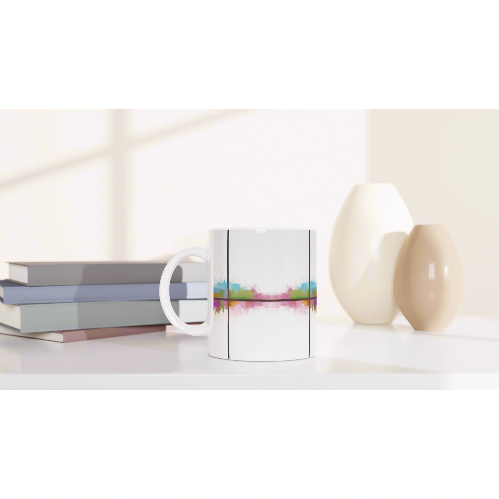White ceramic mug with a multicolored abstract stripe, placed on a desk beside stacked books and beige vases.