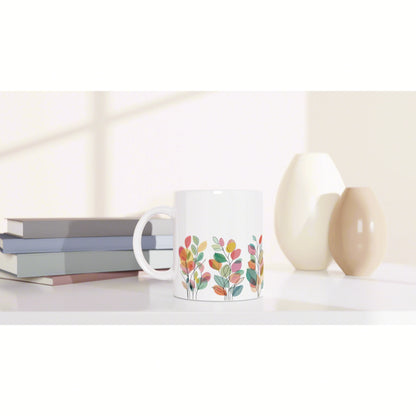 White ceramic mug with a colorful leafy botanical pattern on a white shelf beside stacked books and cream vases.