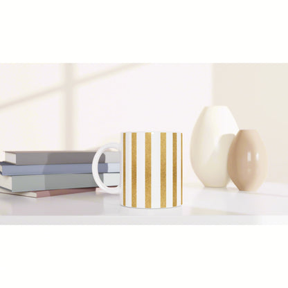 White ceramic mug with gold vertical stripes on a white shelf, beside pastel books and two cream vases.
