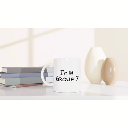 White ceramic mug reading 'I'm in Group 7' on a clean shelf beside books and decorative vases; product mockup.