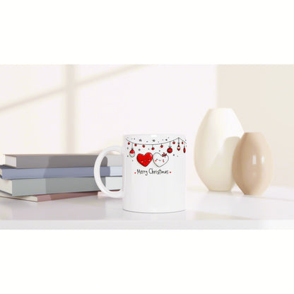 White ceramic mug with a Merry Christmas design featuring red hearts and hanging string lights, beside books and vases.