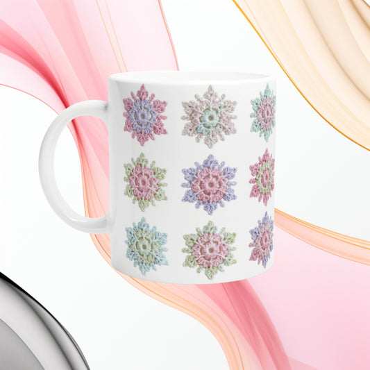 White ceramic mug with pastel crochet-style floral motifs on white, set against abstract pink and beige swirls.