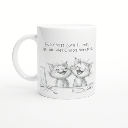 White ceramic mug featuring a pencil sketch of two laughing cats and a German caption about good mood despite chaos.