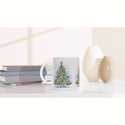 White ceramic mug with colorful watercolor Christmas tree design on a desk beside books and decorative vases.