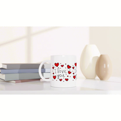 White ceramic mug with red heart motifs and the words I love you, shown on a desk beside stacked books and decorative vases.