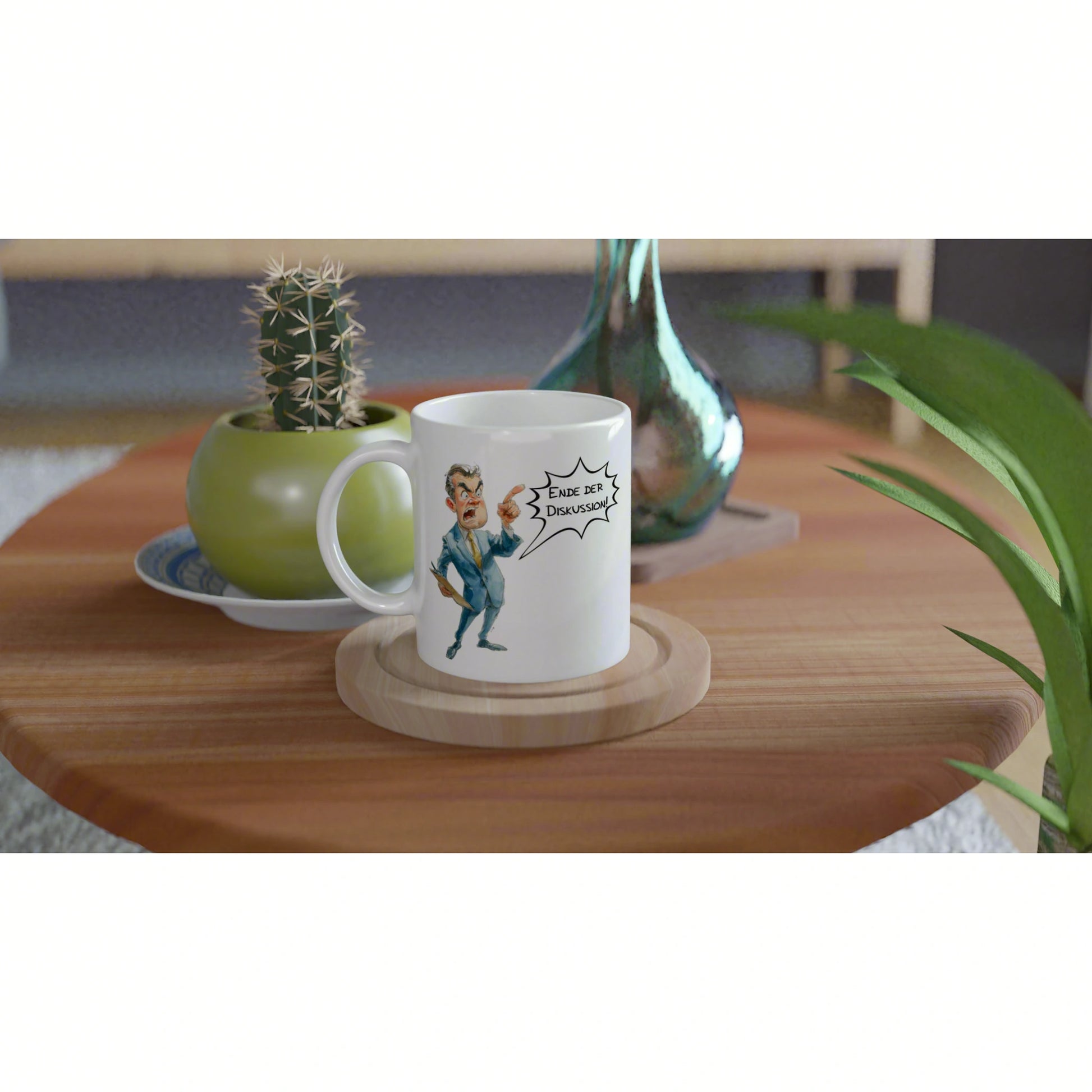 White ceramic mug on a wooden coaster features a cartoon man shouting Ende der Diskussion on a table with a cactus plant.