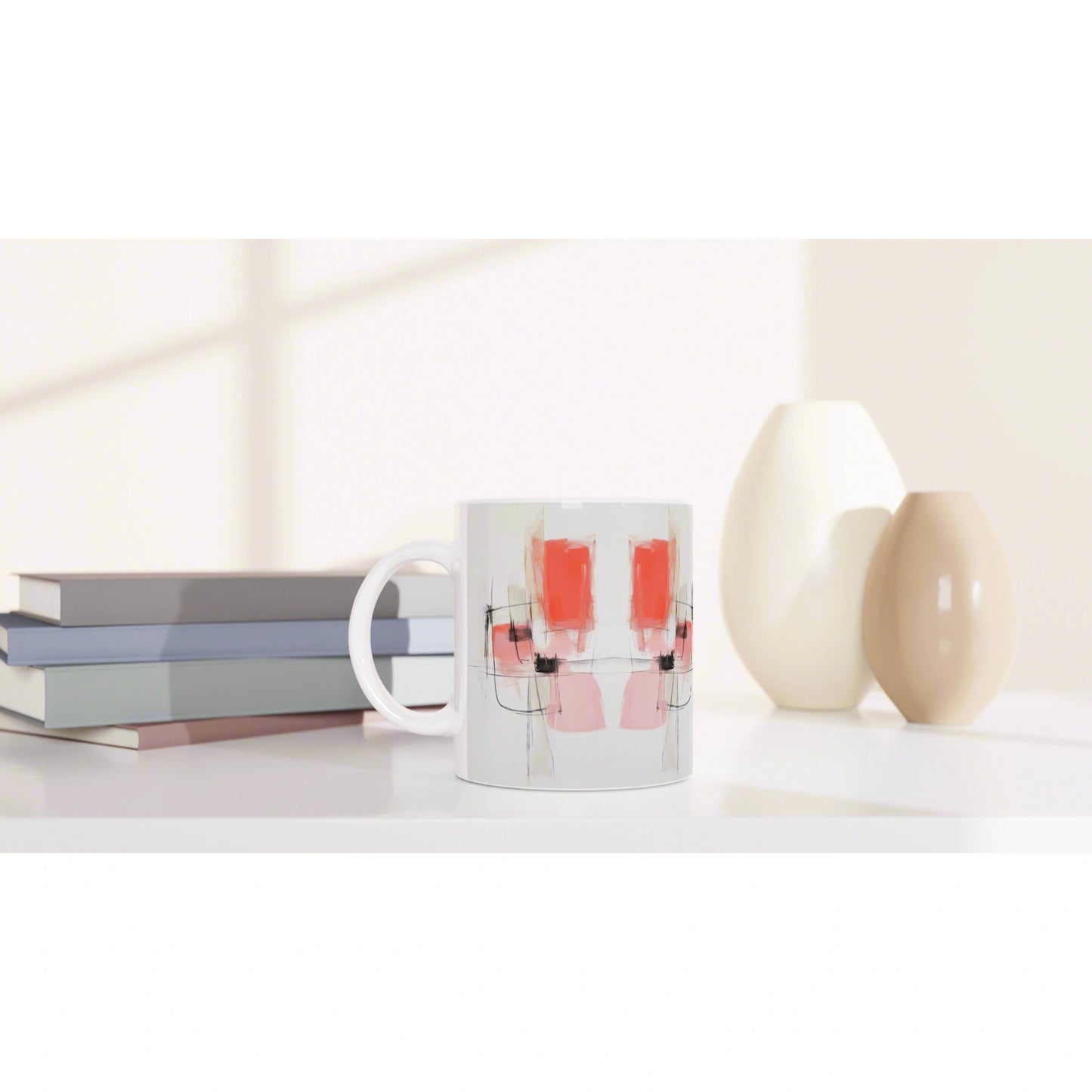 White ceramic mug with an abstract pink and red pattern and black lines, on a desk beside stacked books and cream vases.
