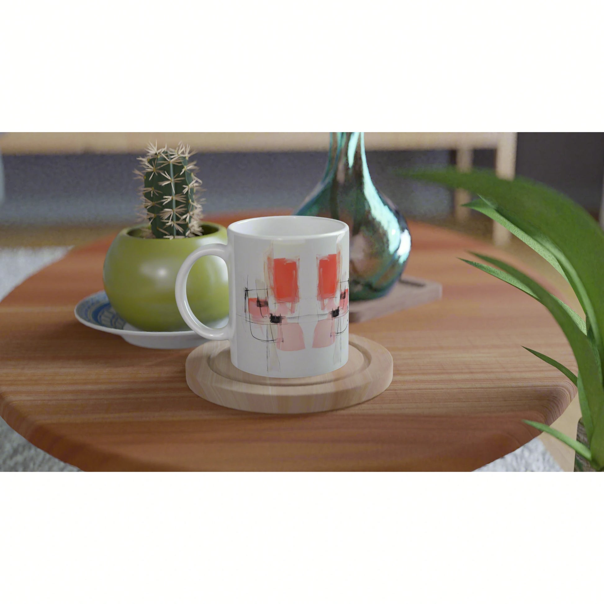 White mug with abstract pink shapes on a wooden coaster, on a wood table beside a green cactus pot and a teal vase.
