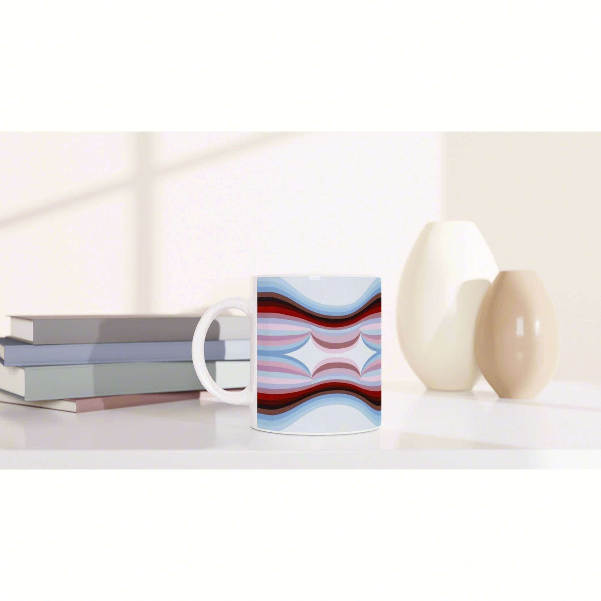 White mug with an abstract blue, pink, and red wave pattern, set on a desk beside stacked books and two ceramic vases.