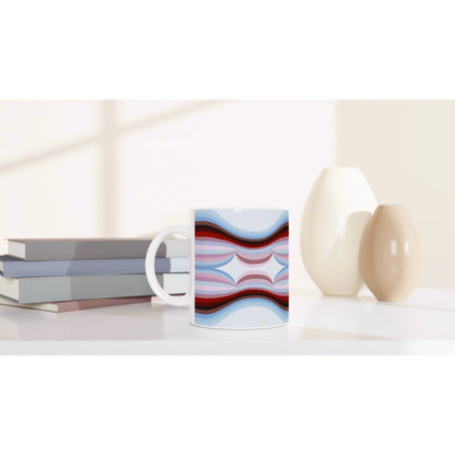 White mug with an abstract blue, pink, and red wave pattern, set on a desk beside stacked books and two ceramic vases.