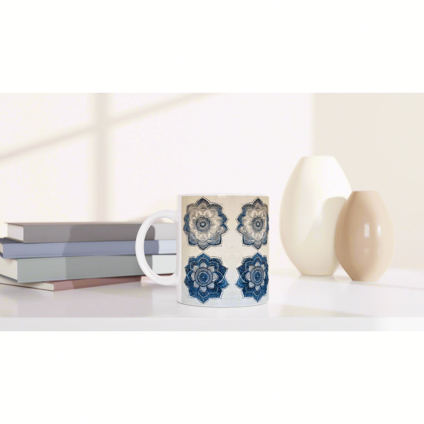 White mug with blue mandala pattern sits on a white desk beside stacked books and beige ceramic vases; a product mockup.