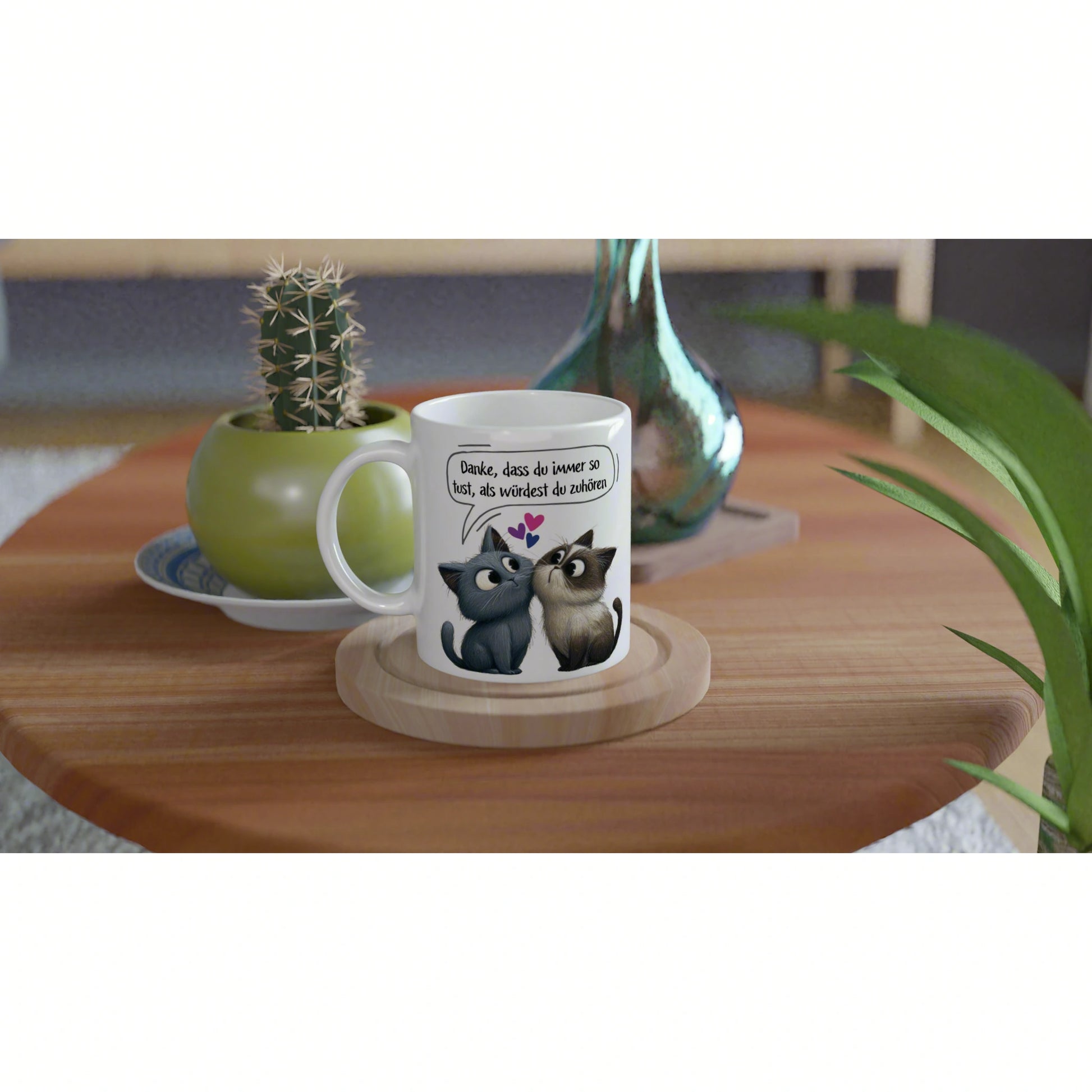 White mug featuring two cartoon cats with a German speech bubble, set on a wooden table with a potted plant and decorative vase.