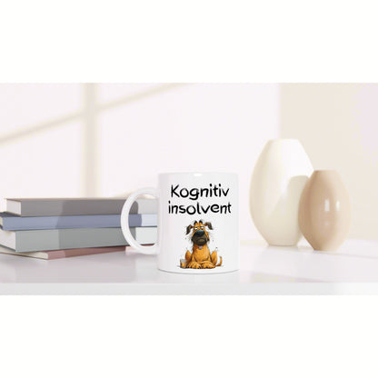 White mug with a cartoon dog and the text Kognitiv insolvent, on a desk with stacked books and decorative vases.