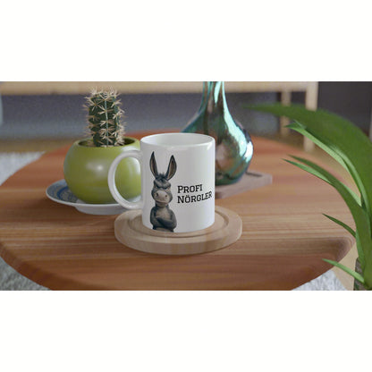 White mug featuring a cartoon donkey with arms crossed and the text 'Profi Nörgler', placed on a wooden coaster amid plants.