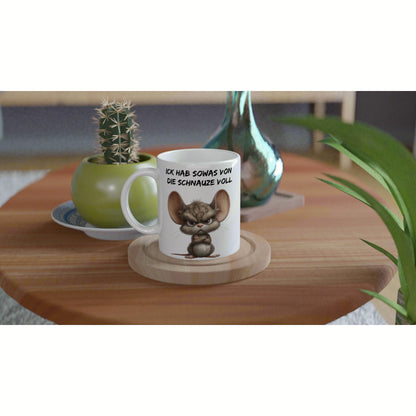 White mug with a cartoon mouse and the slogan ICK HAB SOWAS VON DIE SCHNAUZE VOLL, on a wooden coaster with plants in the background.