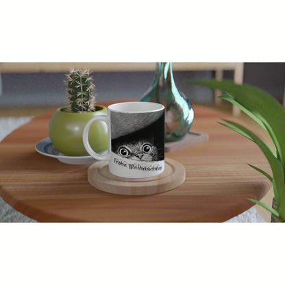 White mug featuring a black-and-white cat peeking from a shadow with holiday text, placed on a wooden table among plants.