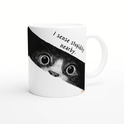 White ceramic mug with a black-and-white cat peeking from the edge and the caption 'I sense stupidity nearby'.