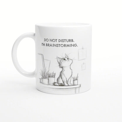 White mug with a pencil-sketch cat on a desk, plants nearby, and the slogan DO NOT DISTURB. I'M BRAINSTORMING.