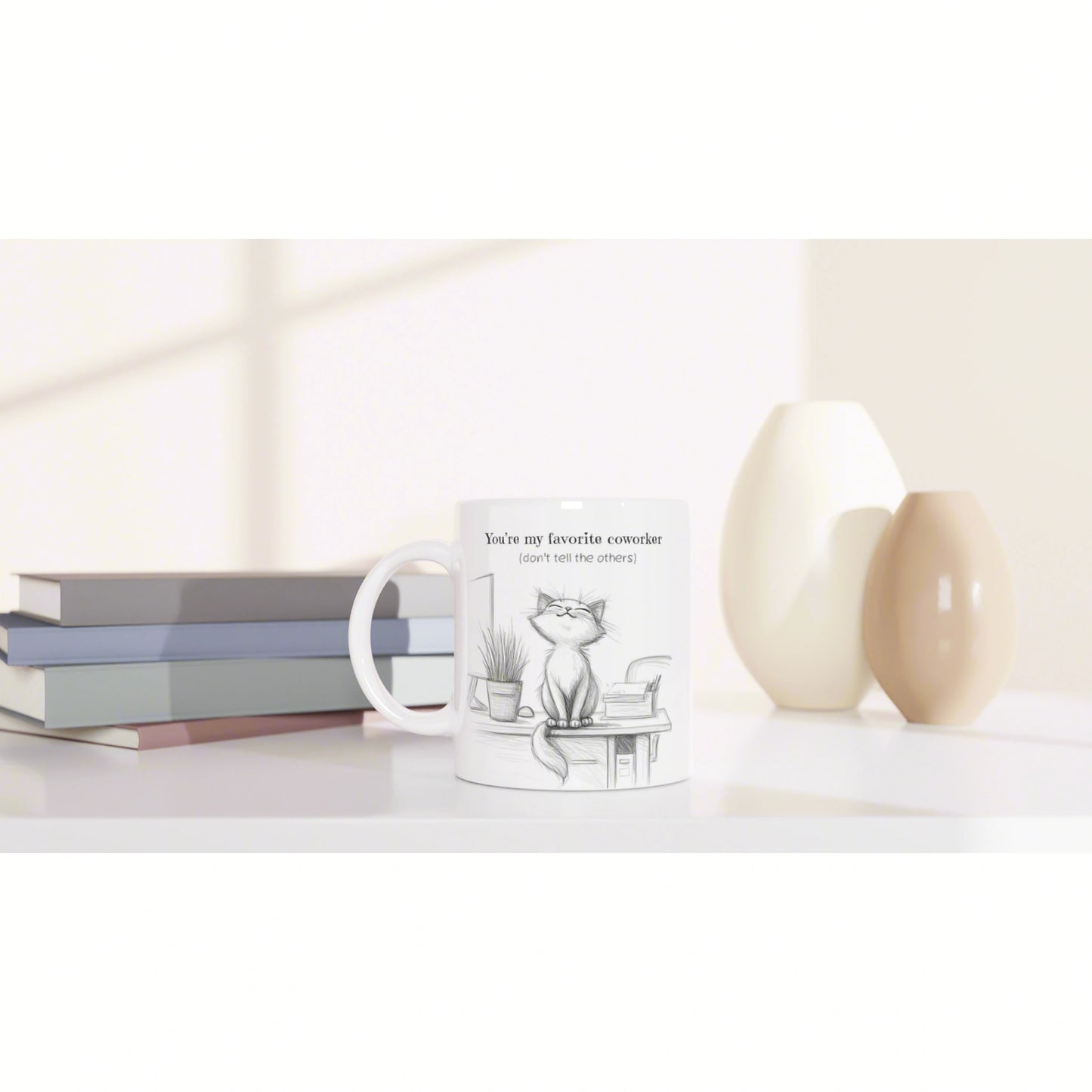 White mug with a cat sketch and caption 'You're my favorite coworker (don't tell the others)' on a desk with books and vases.