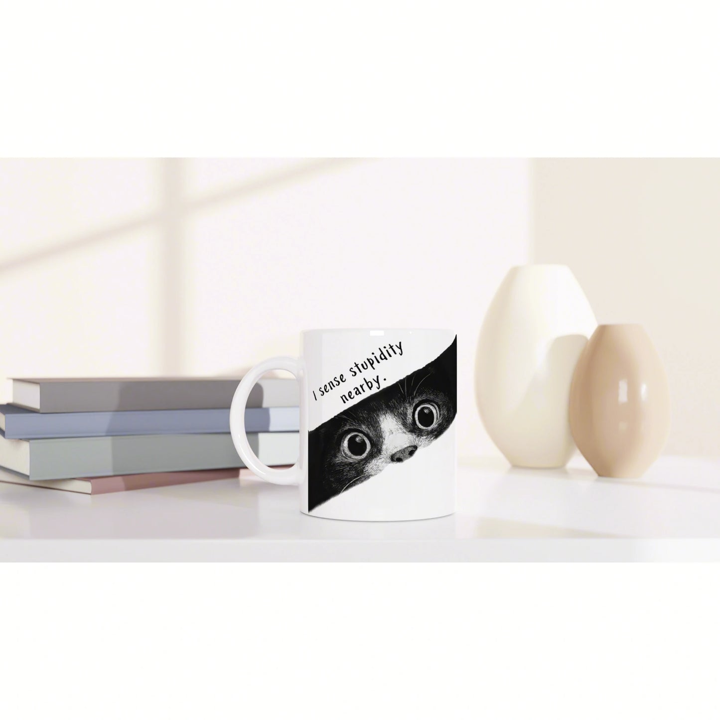 White mug with a black-and-white cat face and the caption 'I sense stupidity nearby,' placed on a desk beside books and decorative vases.