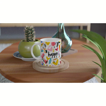 White mug with colorful abstract hearts and the word 'happy' on a wooden coaster, set on a table with plants.