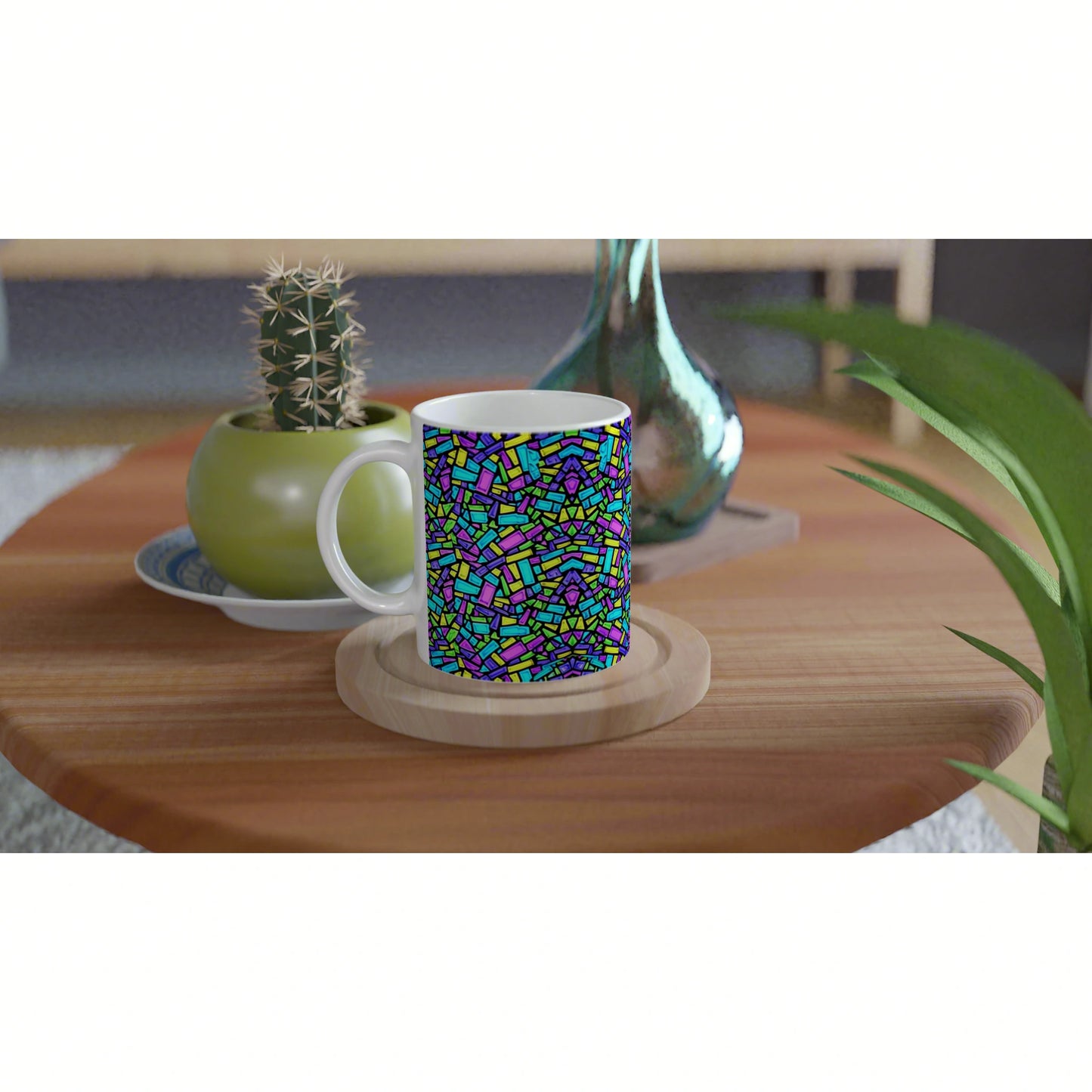 White mug with a vivid mosaic pattern on a wooden coaster, on a wooden table beside a cactus and a decorative vase.