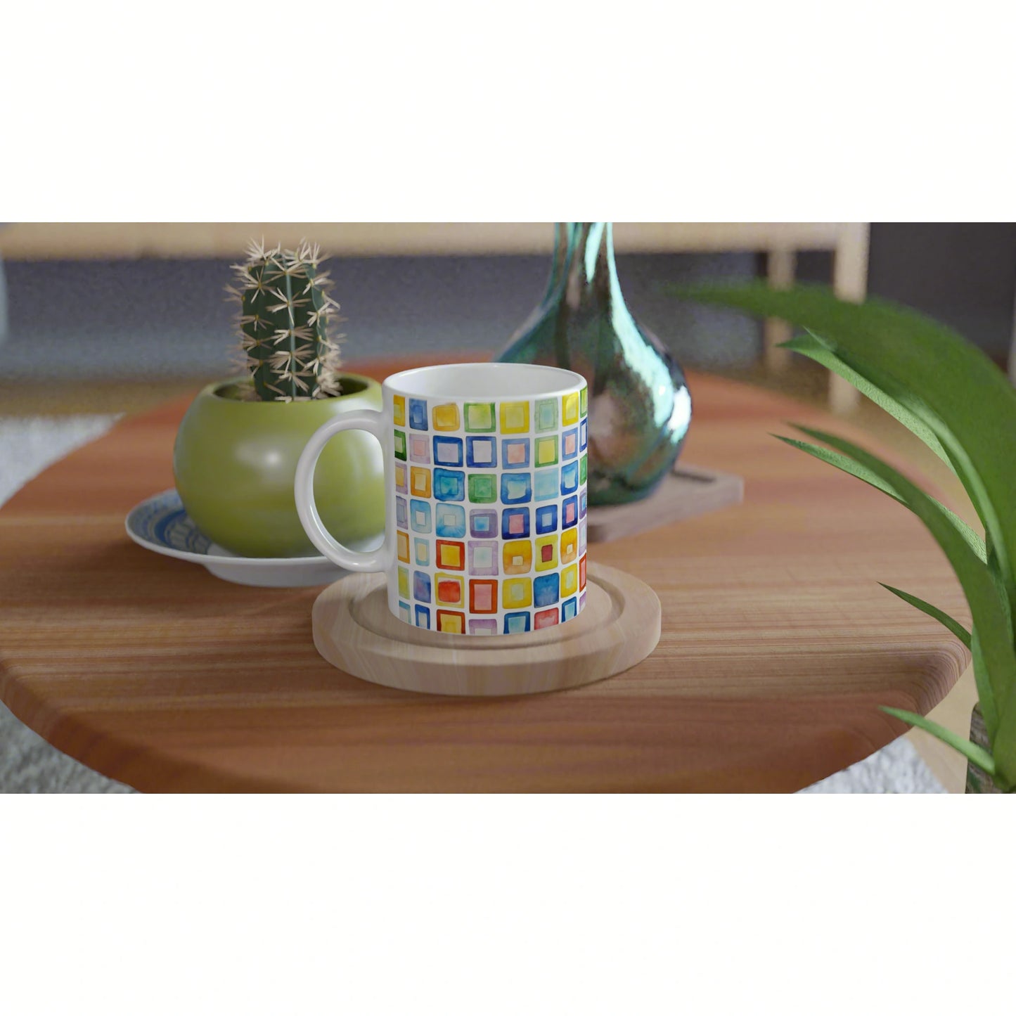 Product image: white mug with a colorful square mosaic pattern on a wooden table, flanked by a green cactus pot and a glass vase.