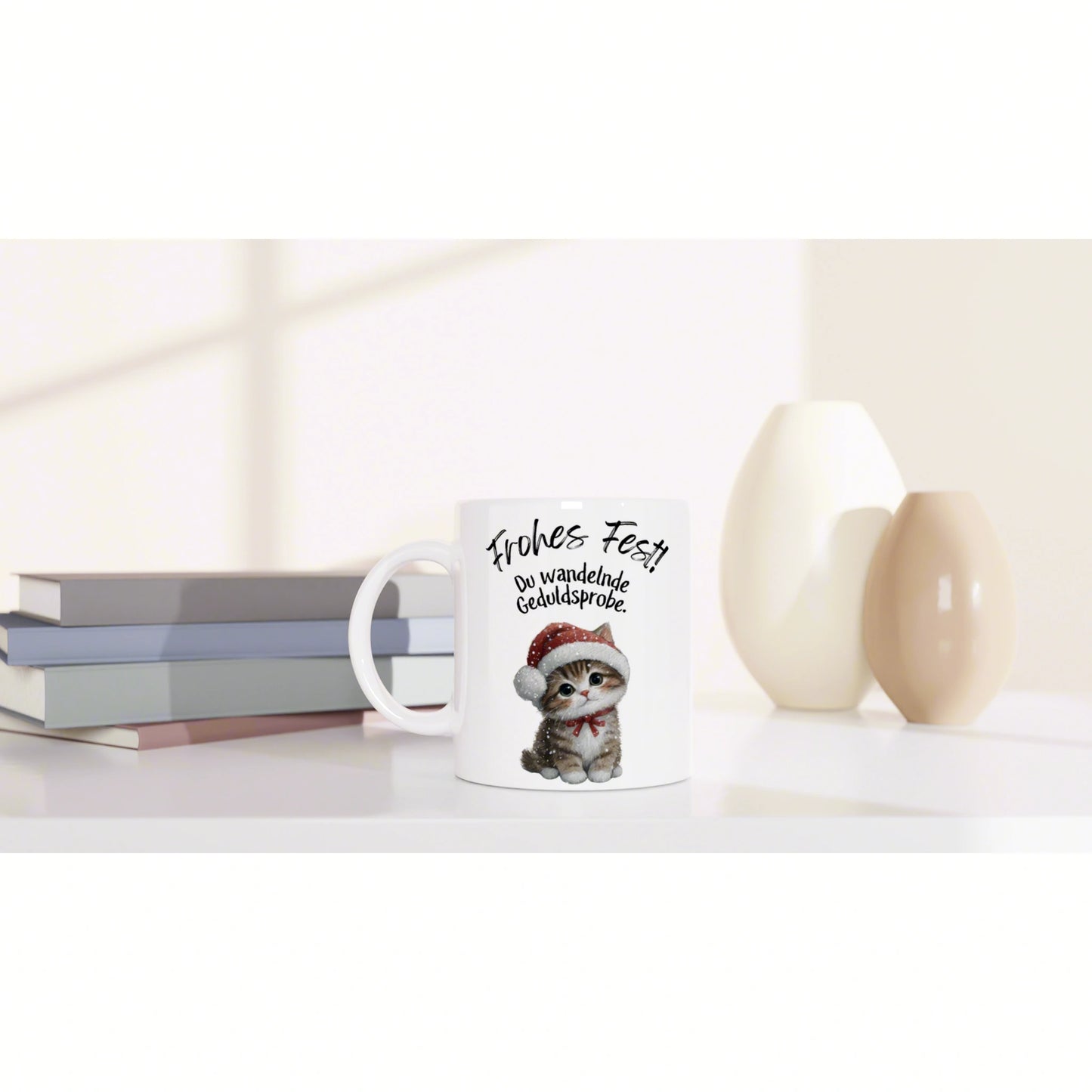 White mug with a cute cat in a Santa hat and the German text 'Frohes Fest! Du wandelnde Geduldsprobe.', on a shelf with books and decorative vases.