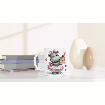 White mug featuring a cute hippo in a pink dress holding a cake, on a desk with books and vases - product mockup.