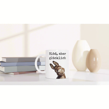 White mug featuring a cartoon donkey and the text 'Blöd, aber glücklich', on a desk with books and pale vases.