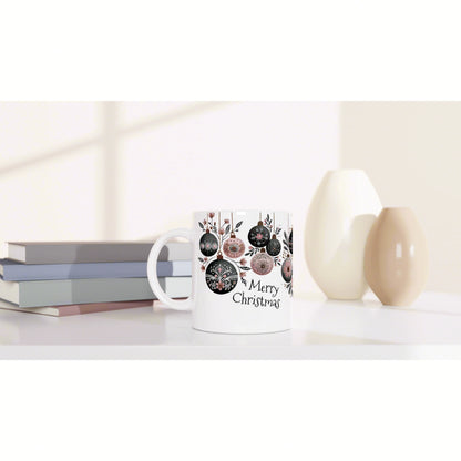 White mug featuring decorative ornaments and 'Merry Christmas' text, placed beside books and ceramic vases in a soft studio.