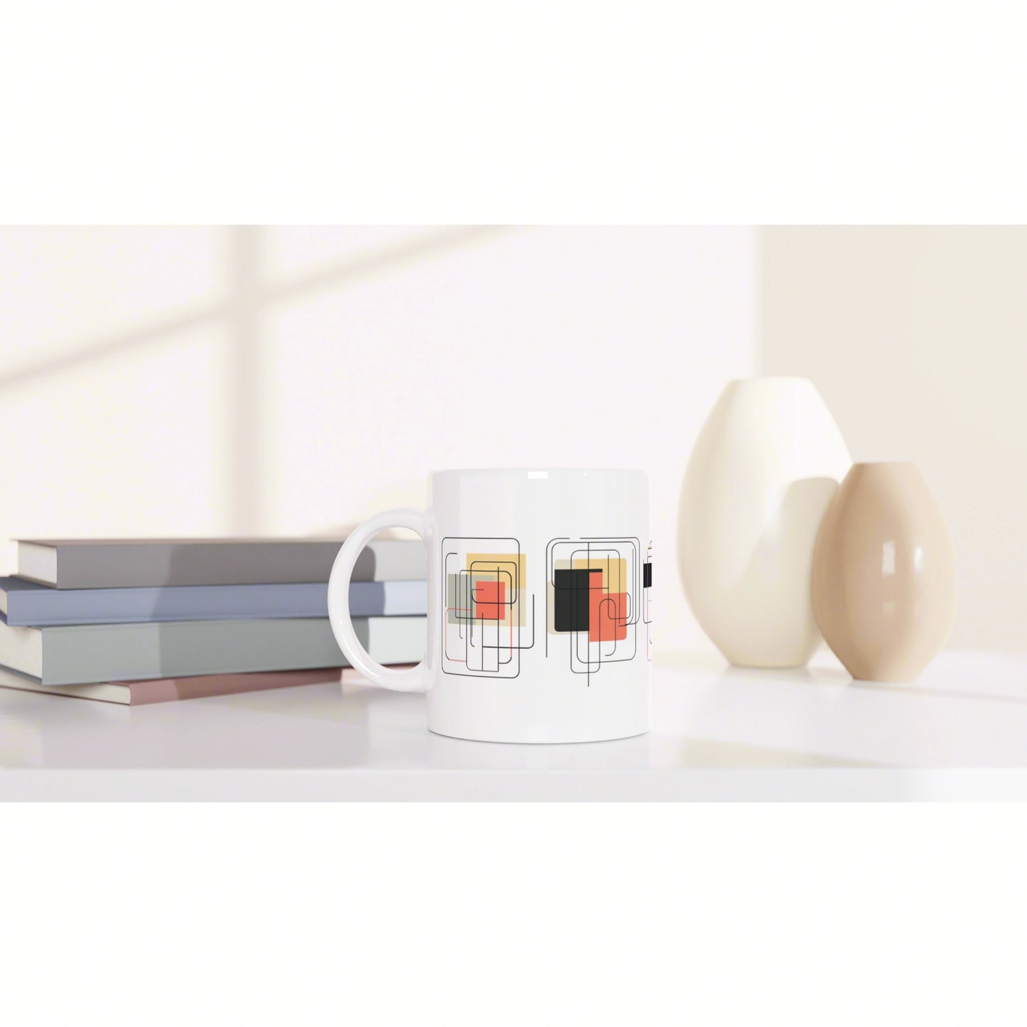 White ceramic mug with overlapping square-pattern design in black and colored blocks, beside books and beige vases.