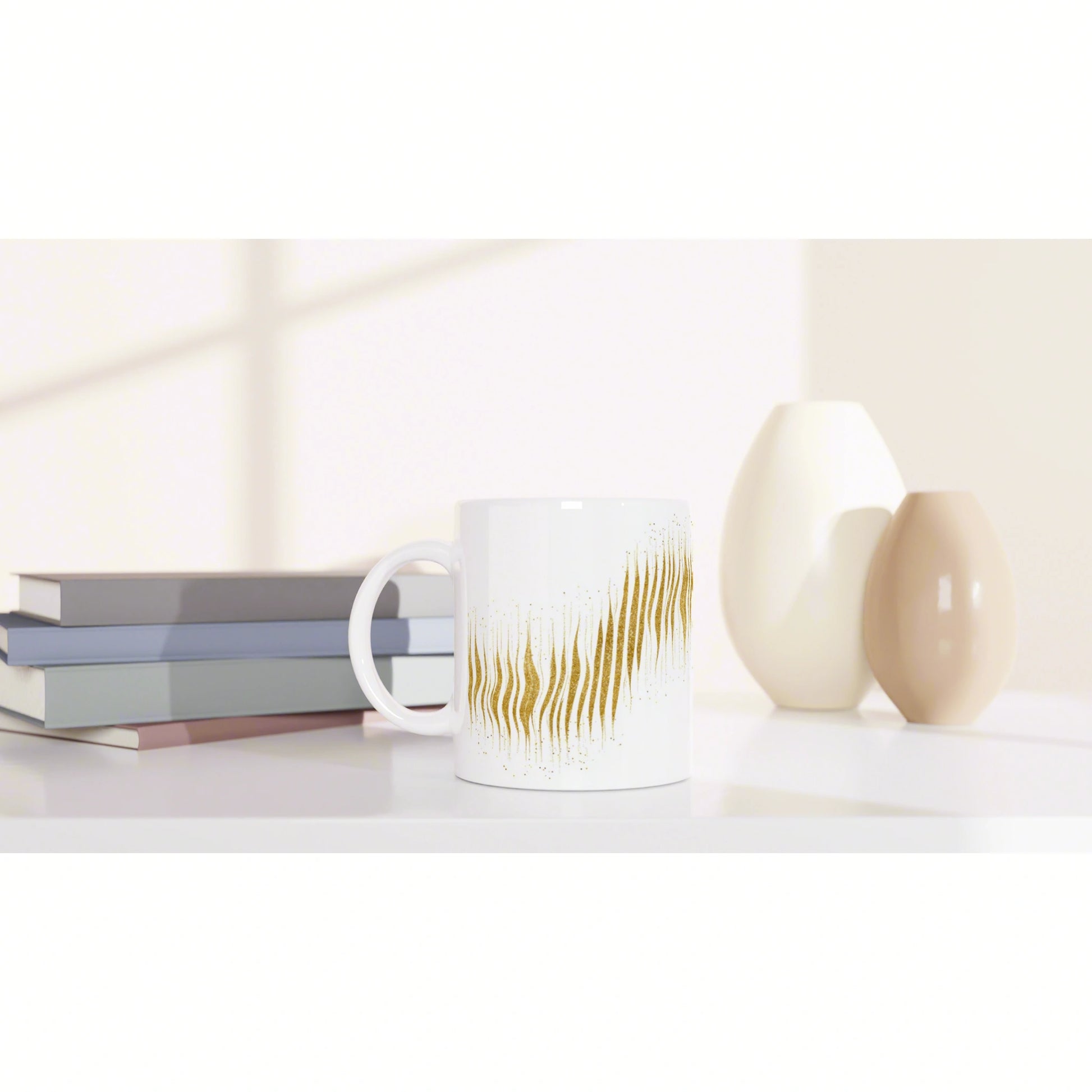 White mug with a gold abstract flame pattern on a desk beside stacked books and pale decorative vases.