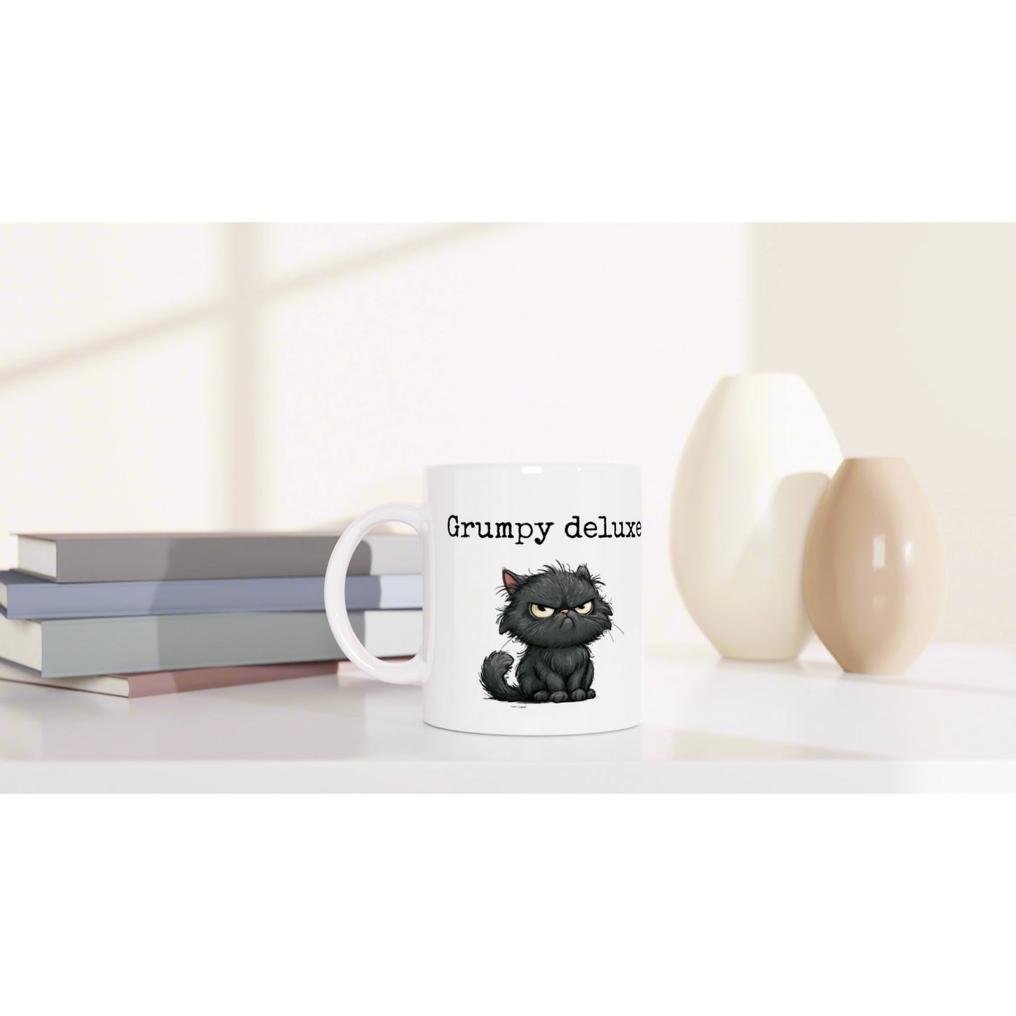 White mug featuring a grumpy black cat illustration and the text 'Grumpy deluxe', placed beside stacked books and decorative vases.