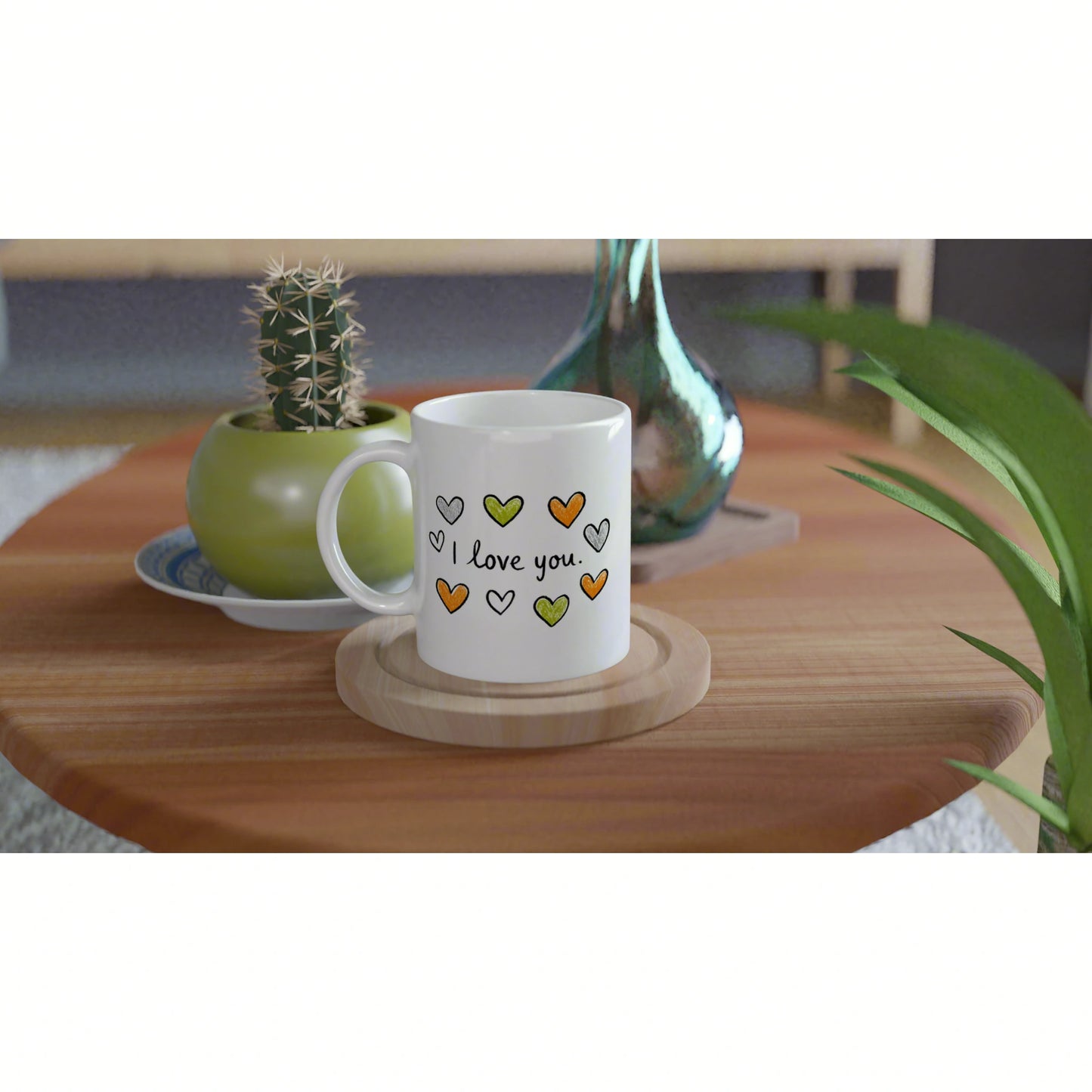 White mug with colorful hearts and 'I love you' on a wooden coaster, on a table with a cactus and a decorative vase.