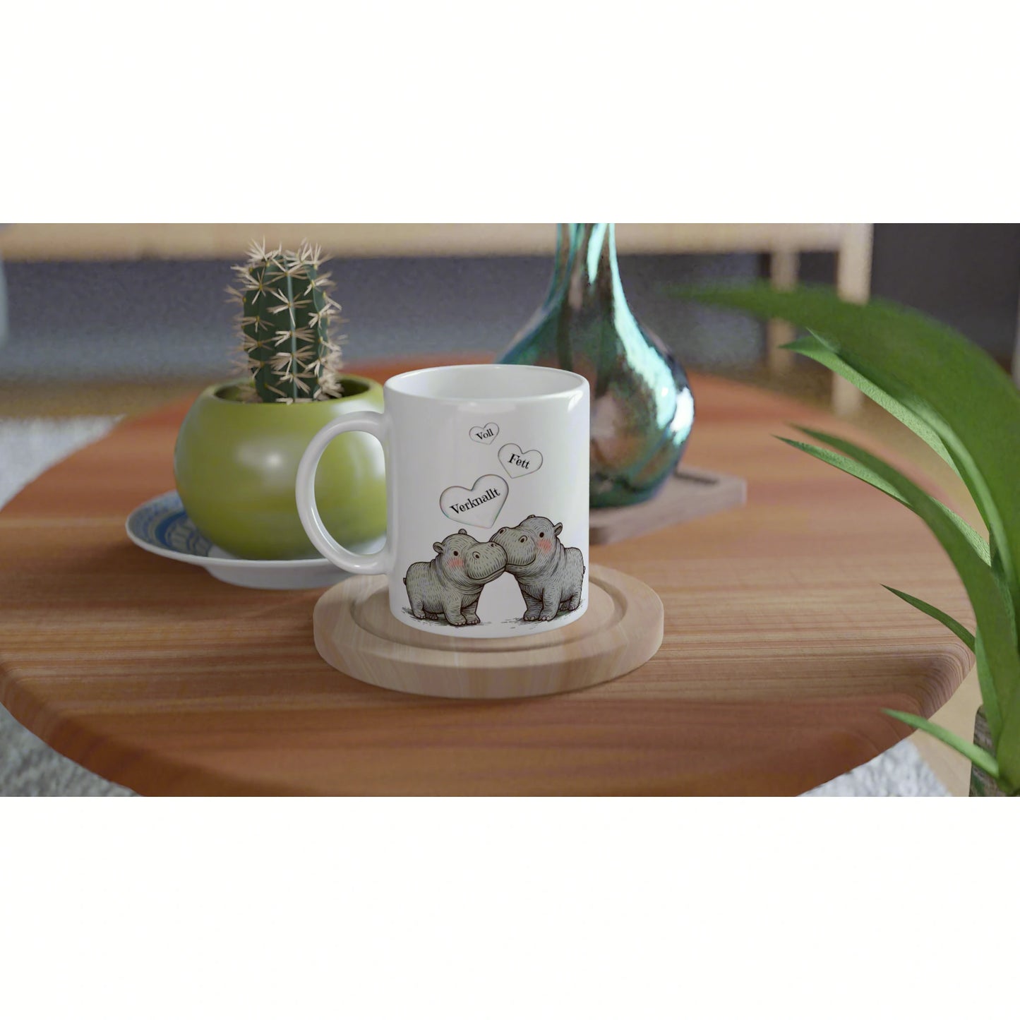 White mug with two cartoon hippos and heart speech bubbles, on a wooden coaster near a potted cactus.